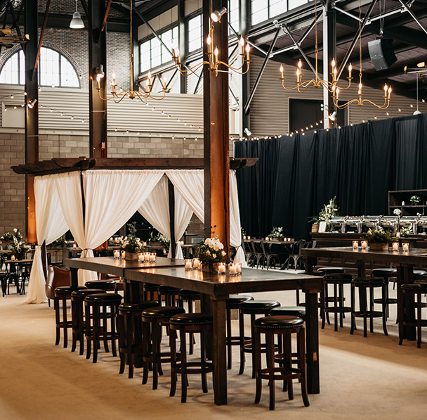 Wedding reception area with bar seating, high wooden tables, and curtained lounge area inside industrial building