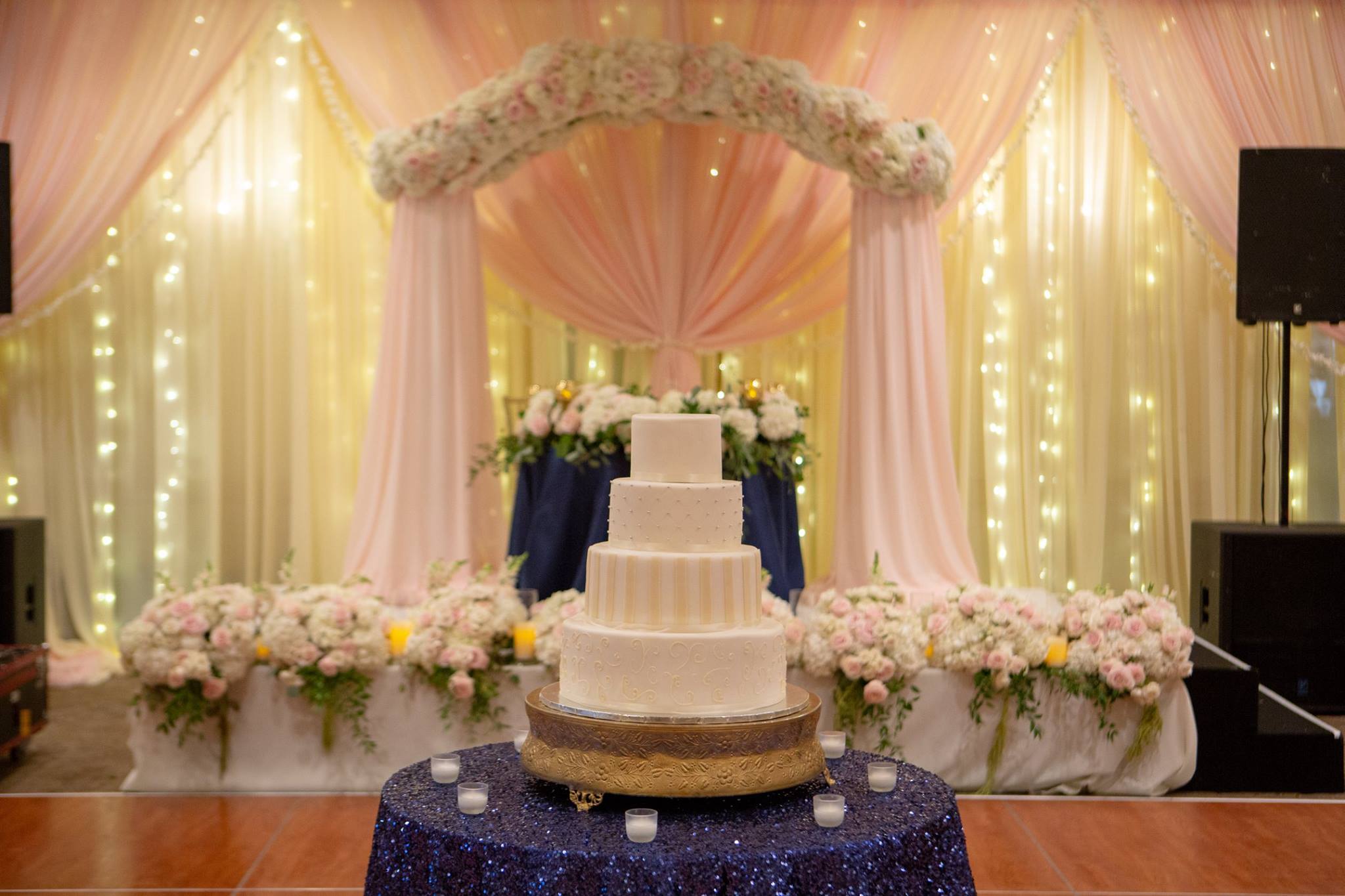 Staged four-tier wedding cake in front of rose and white pipe and drape setup at wedding