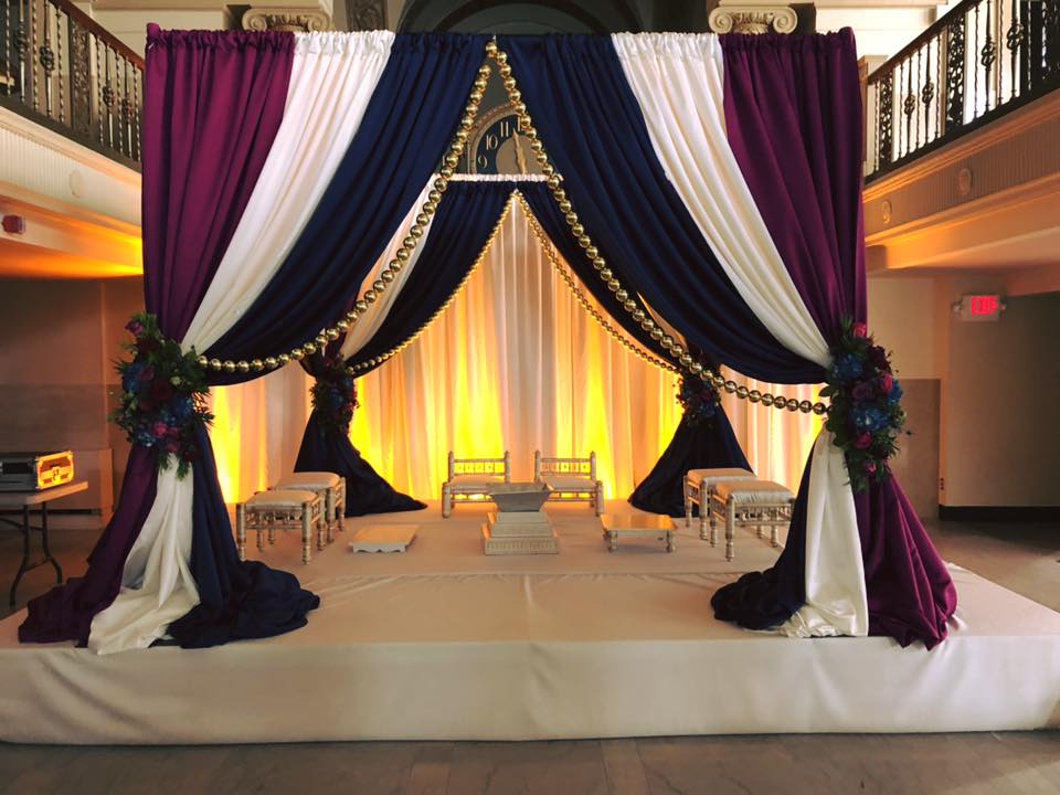 Pipe and drape set in royal purple, white, and navy blue, with flower accents and golden curtain pulls around white sitting furniture